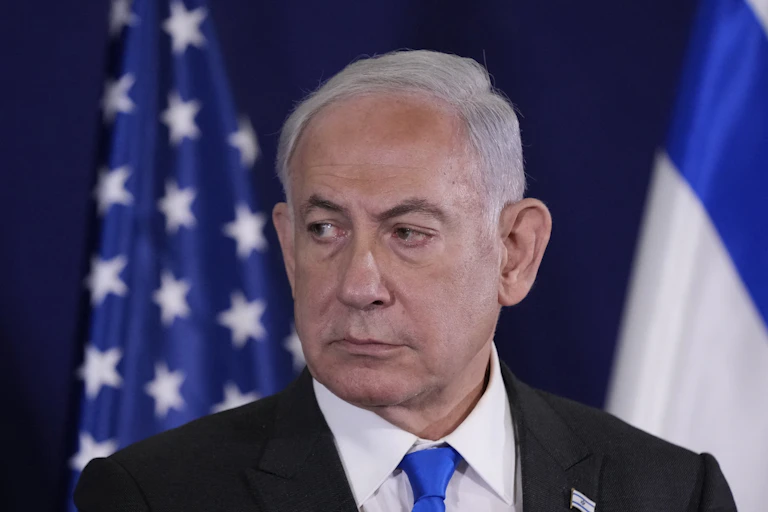 A white-haired man looking solemn in front of two flags.