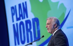 A grey-haired bearded man speaks into a microphone with the blue-and-green logo for Plan Nord beside him.