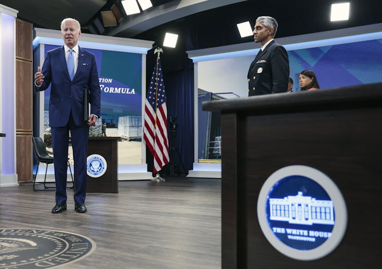 Two men in suits stand in a room with screens and flags.