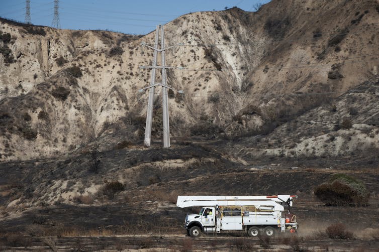 A white utility truck drives toward a transformer tower framed by hills.