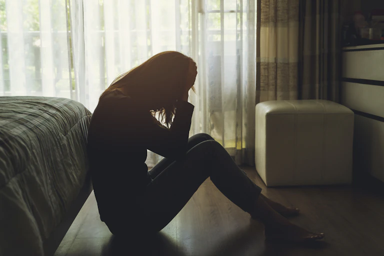 A woman sitting on the floor against a bed, appears stressed or depressed.