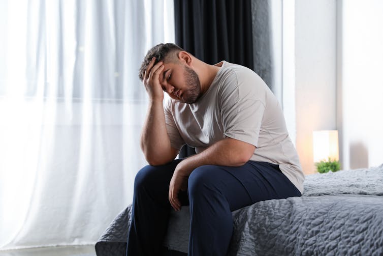 A man sits on a bed, appears exhausted.