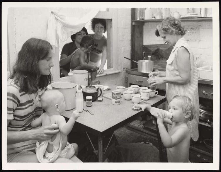 A gorup of women and toddlers crowd around a kitchen table in the 1970s.