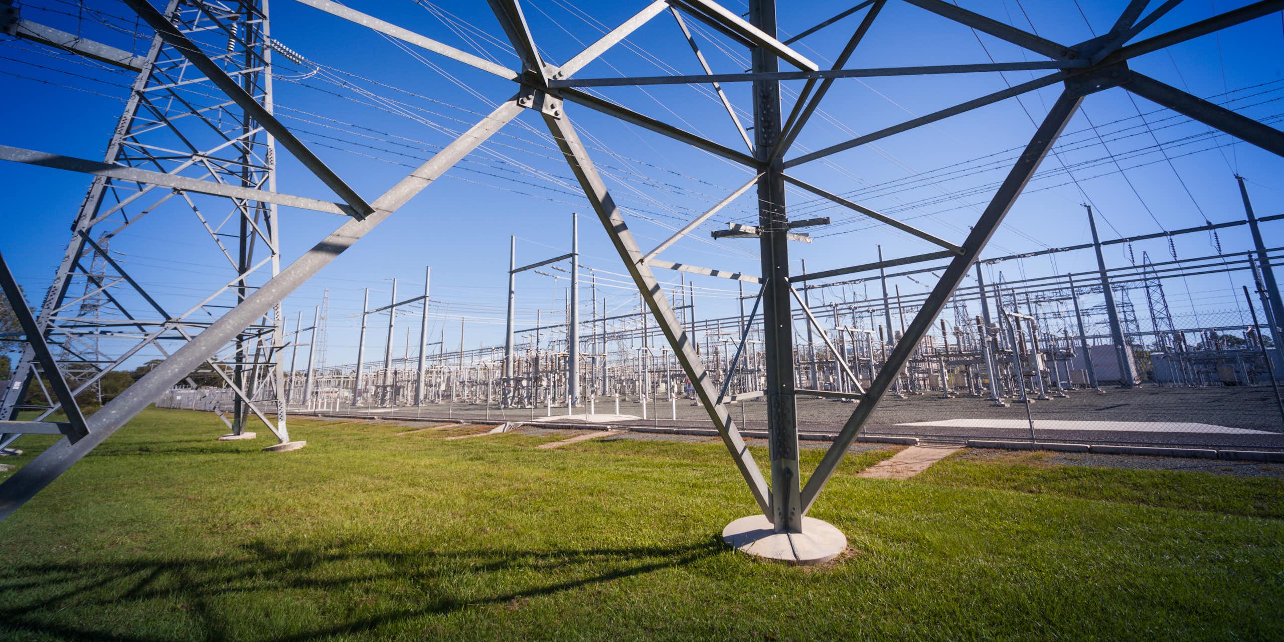 Peering through the metal structure of an electricity pylon into a power substation in Queensland, standing on green grass with a blue sky in the background