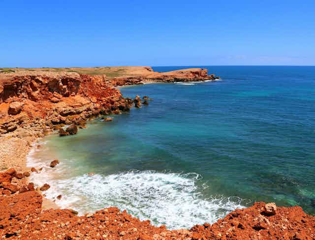 A red rocky ocean coast with blue skies and teal water.
