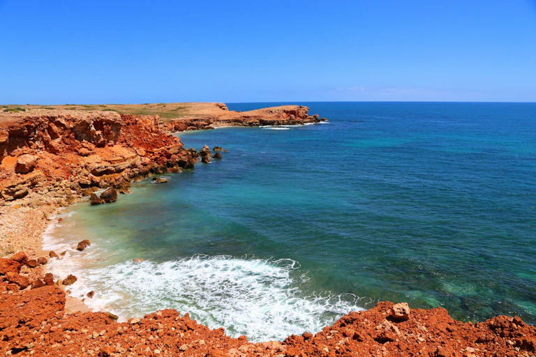 A red rocky ocean coast with blue skies and teal water.