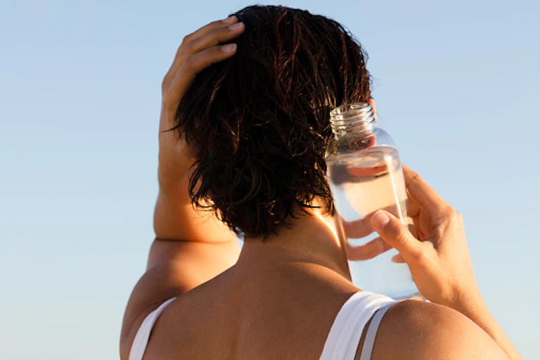 Back view of a woman holding a bottle of water to her neck. with her other hand on her head