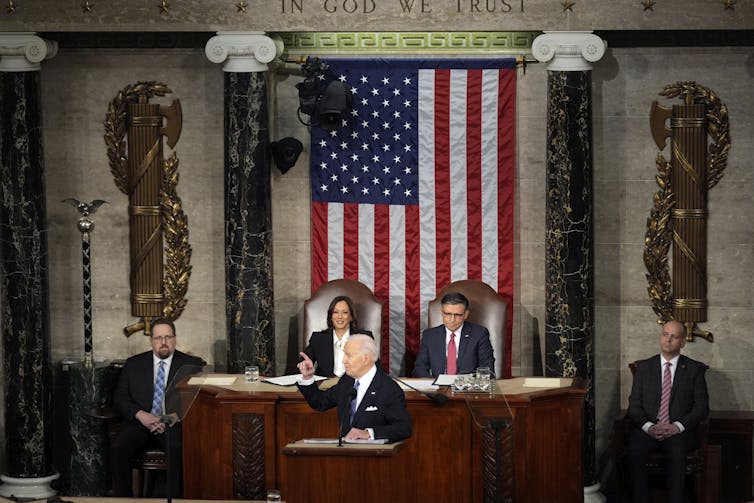 Joe Biden is seen standing at a podium, in front of a large American flag and several people around him, including Vice President Kamala Harris