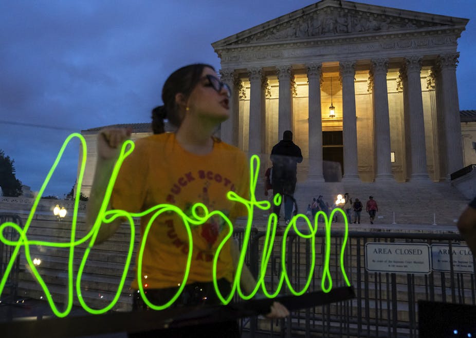 A young woman with brown hair, glasses and a yellow shirt holds up a neon green sign that says 'abortion' outside the Supreme Court building on an evening with a dark blue sky.