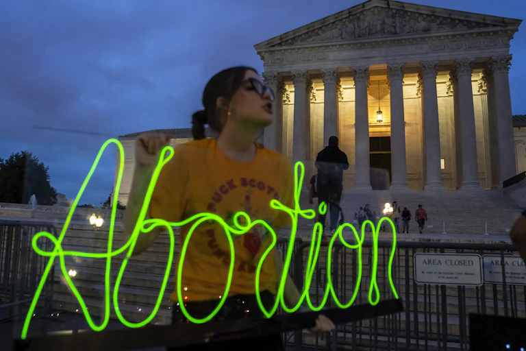 A young woman with brown hair, glasses and a yellow shirt holds up a neon green sign that says 'abortion' outside the Supreme Court building on an evening with a dark blue sky.