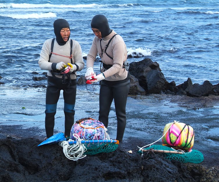 Dos buceadoras hablan de pie sobre una roca con el mar detrás.