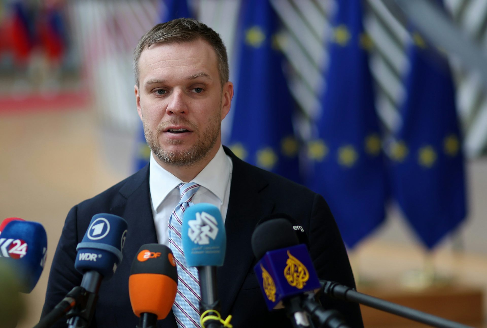 Gabrielius Landsbergis speaking into multiple microphones at a press conference with European flags in the background. 