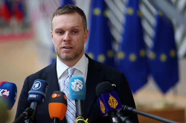 Gabrielius Landsbergis speaking into multiple microphones at a press conference with European flags in the background. 