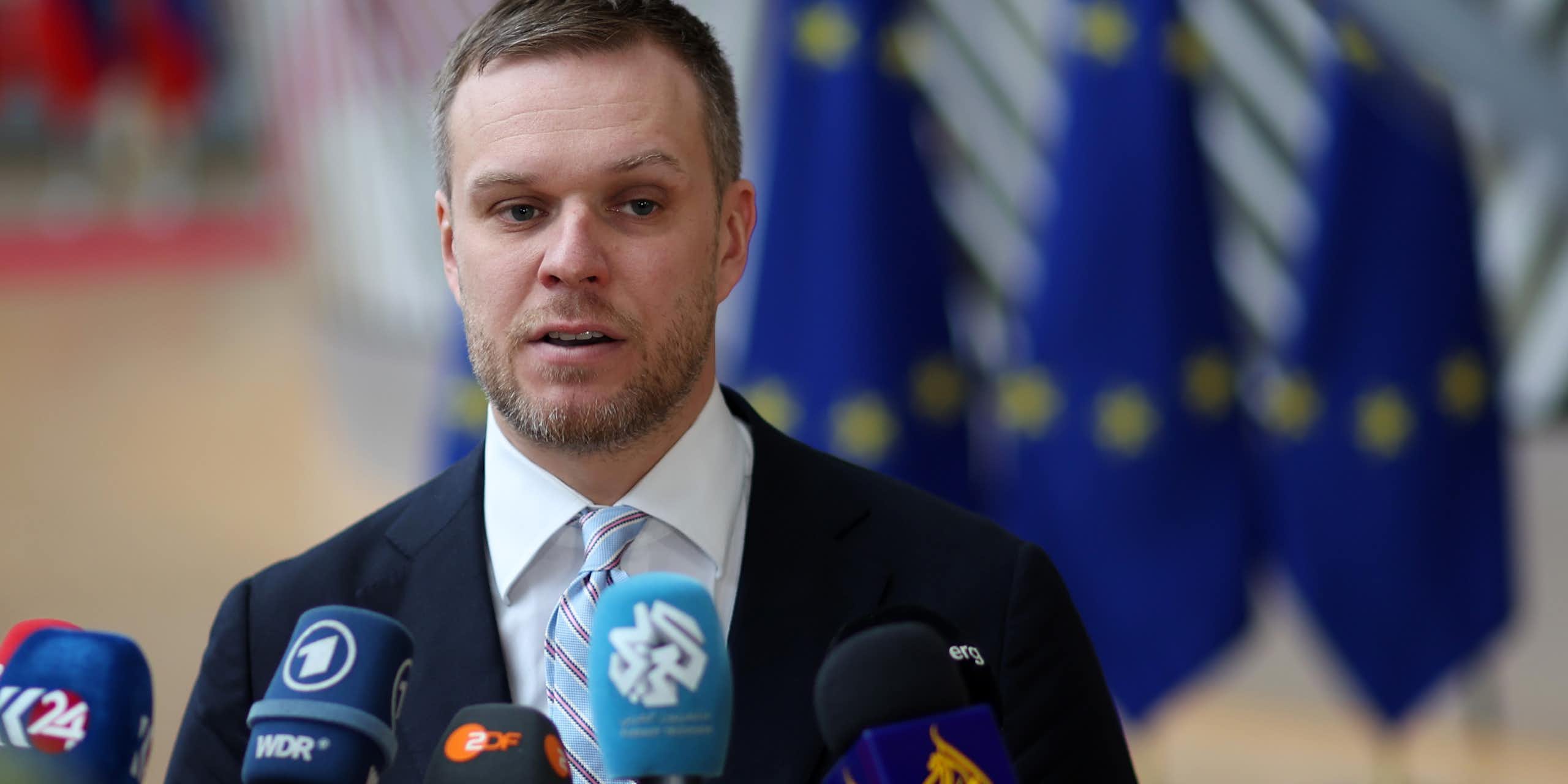 Gabrielius Landsbergis speaking into multiple microphones at a press conference with European flags in the background. 