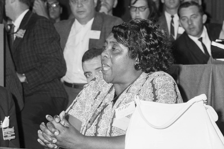 A Black woman sits behind a desk with her hands clasped together as a gathering of white men listen to her speech.at a desk and in speaking with a microphone.