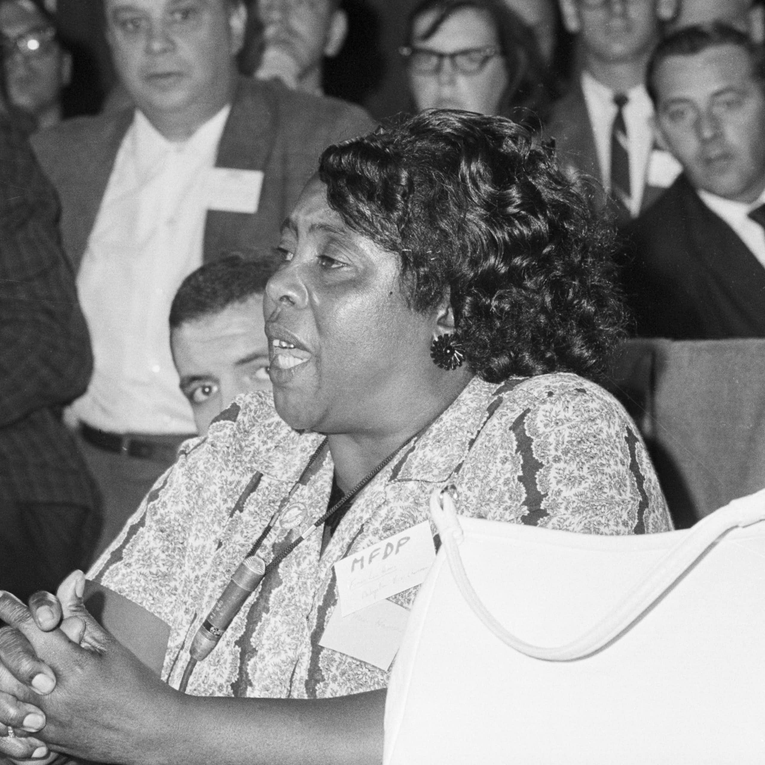 A Black woman sits behind a desk with her hands clasped together as a gathering of white men listen to her speech.at a desk and in speaking with a microphone. 