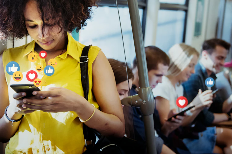 Dans un transport en commun, au premier plan, une jeune femme est sur son smartphone, des dessins d'émoticones positifs, des pouces levés et coeur sortent du smartphone. En arrière-plan, des gens assis sont également sur leurs smartphones dont sortent les mêmes dessins d'émoticones, pouces levés et coeur.