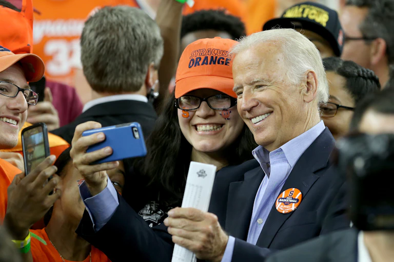 A gray haired man taking a selfie with a woman in an orange baseball cap.