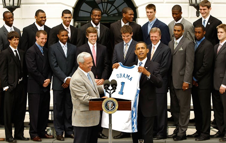 A man holding a team jersey with the name 'Obama' on it, standing in front of a large group of men in suits.