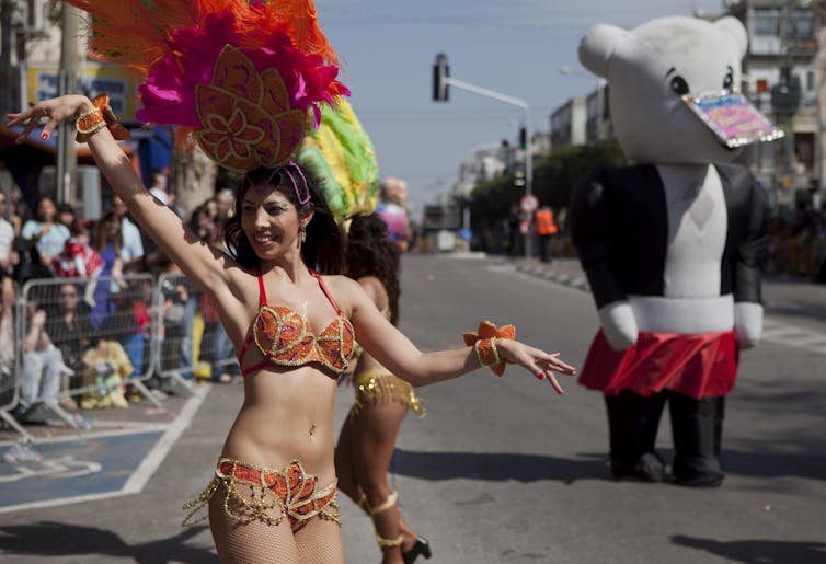 A woman in an orange bikini-style outfit and a large red headdress dances in the street near a tall stuffed bear figure.