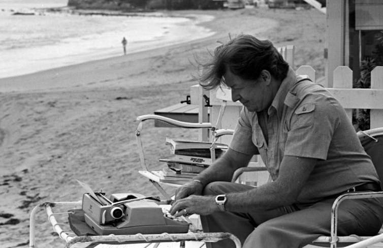 Black and white photo of a middle-aged man sitting at a typewriter by the sea.
