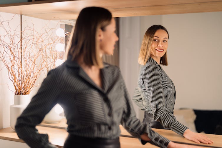 A young woman in a collared shirt looks confidently into a mirror