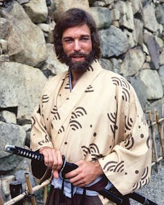 Bearded man with shoulder length brown hair wearing a kimono and holding a samurai sword.