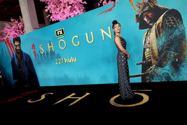 Young Asian woman poses in front of a blue display featuring images from the series 'Shogun.'