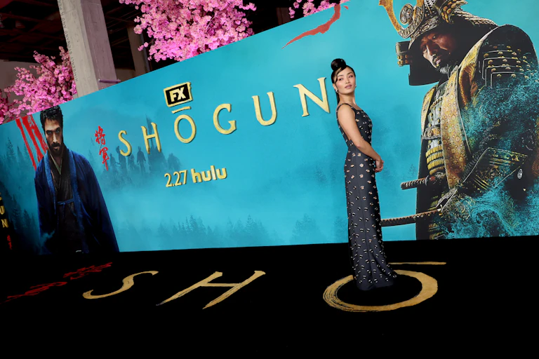 Young Asian woman poses in front of a blue display featuring images from the series 'Shogun.'