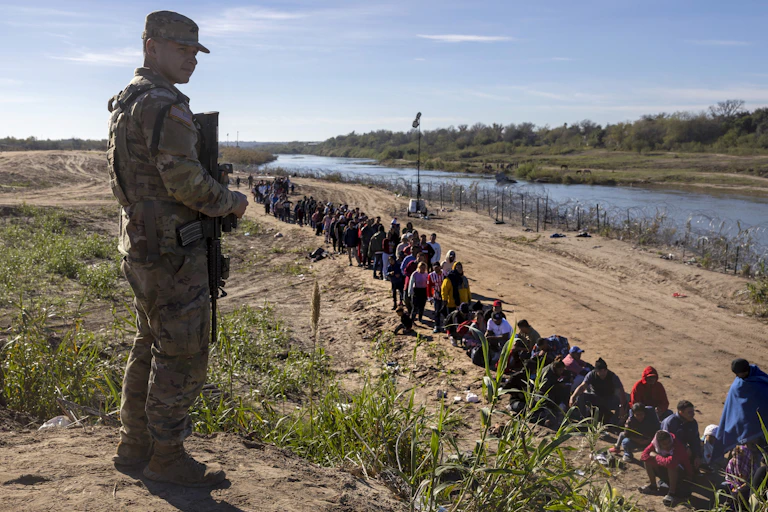 A young person in camo uniform holds a gun and stands on a hill overlooking a low row of people who stand near a river.