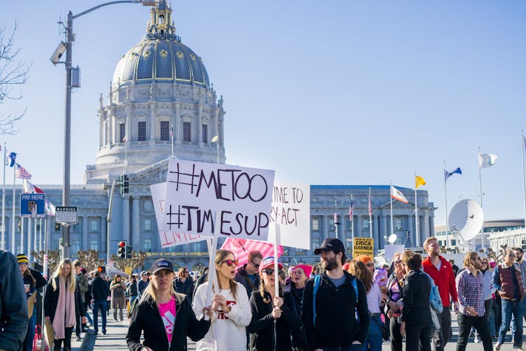 A group of protesters holding signs with #metoo and #timesup slogans written on them in front of a domed government building