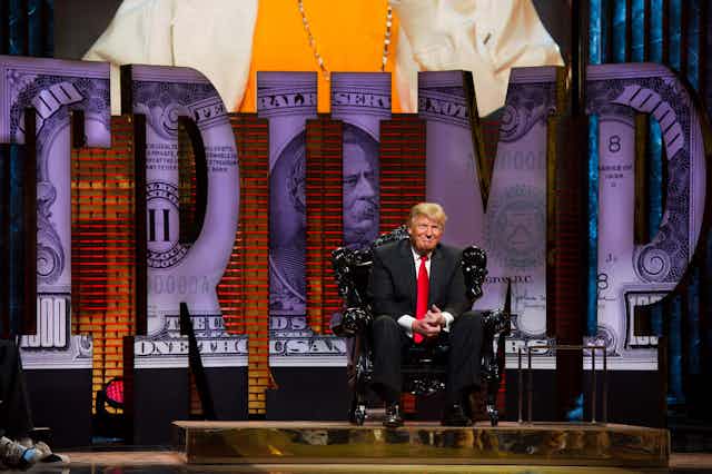 white man with suit and red tie sits on stage in front of big letters that read trump with an image of a 100 dollar bill inside