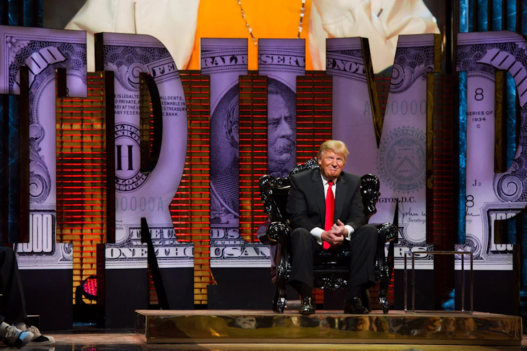 white man with suit and red tie sits on stage in front of big letters that read trump with an image of a 100 dollar bill inside