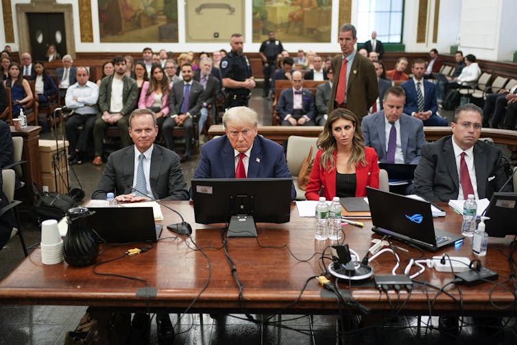 several men and a woman sit at a wooden table in a courtroom with observers behind them