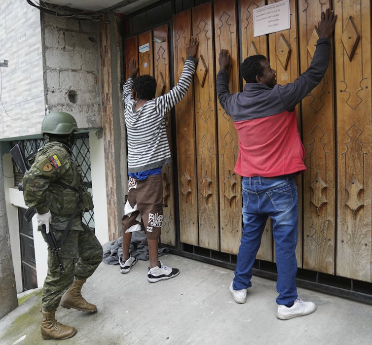 Two men stand with their arms against a wall as an armed soldier stands behind them.