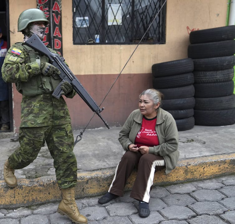 A soldier with his weapon drawn walks past a woman sitting on the curb.