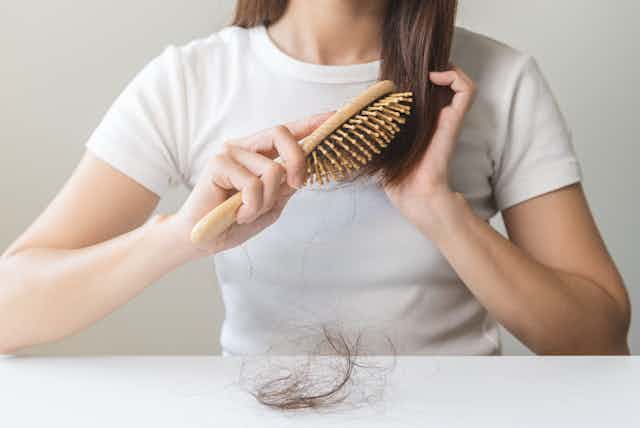 Uma mulher escova o cabelo usando uma escova de madeira. Há um tufo de cabelo sobre a mesa à sua frente.
