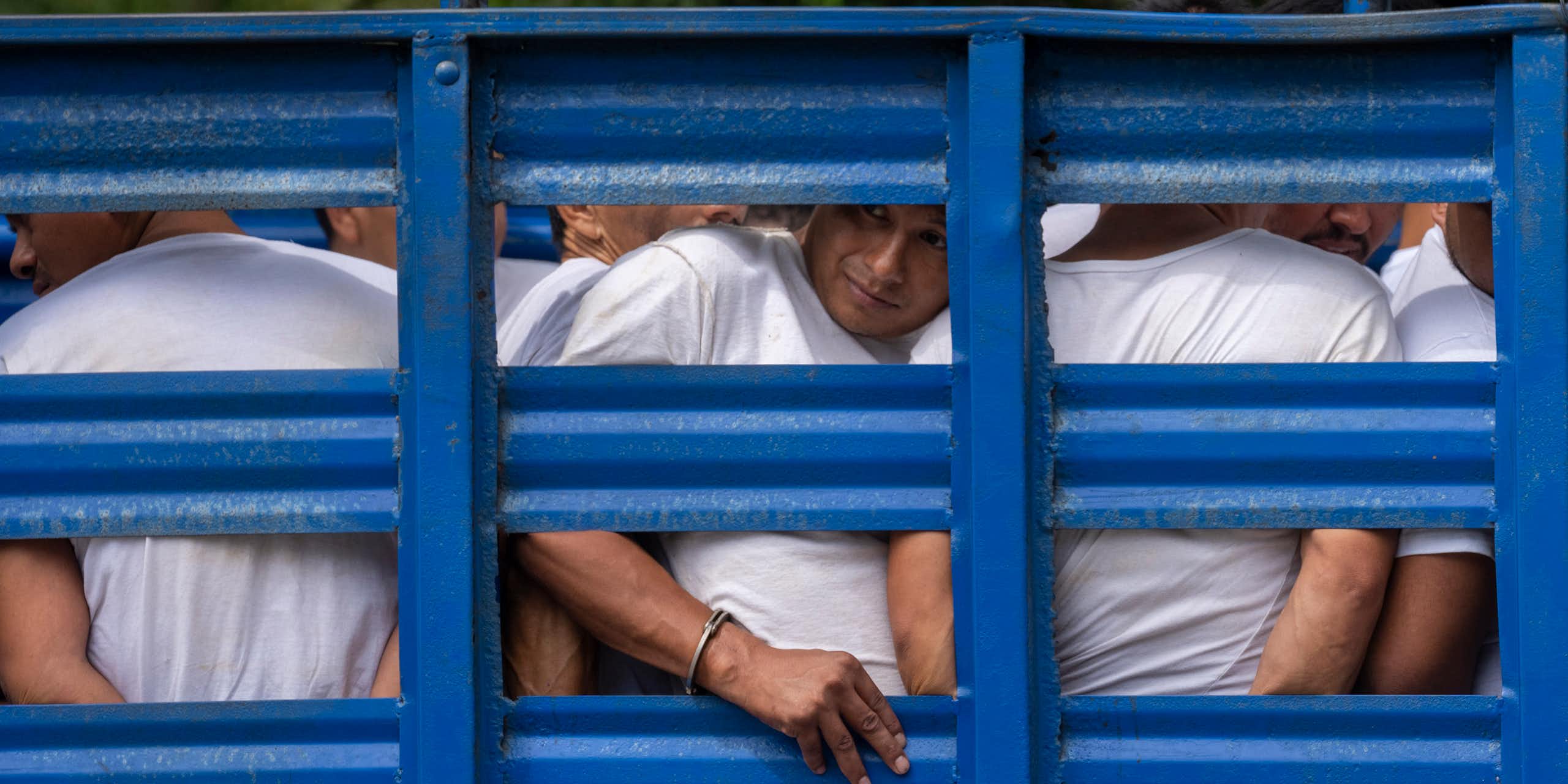 Men with their hands in handcuffs look between the slats of a blue cargo truck.
