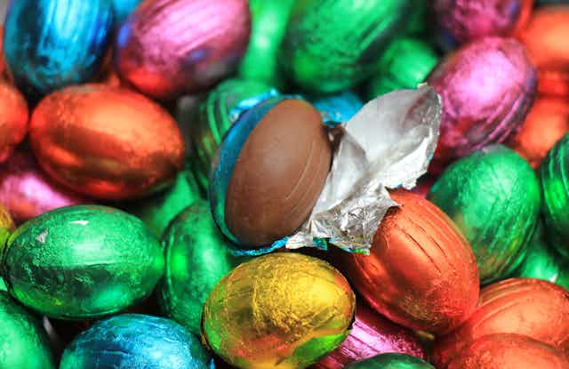 A collection of Easter eggs in brightly coloured foil.  One is unwrapped revealing the chocolate inside.