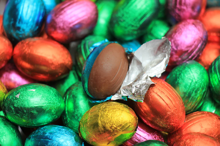 A collection of Easter eggs in brightly coloured foil. One is unwrapped revealing the chocolate inside.