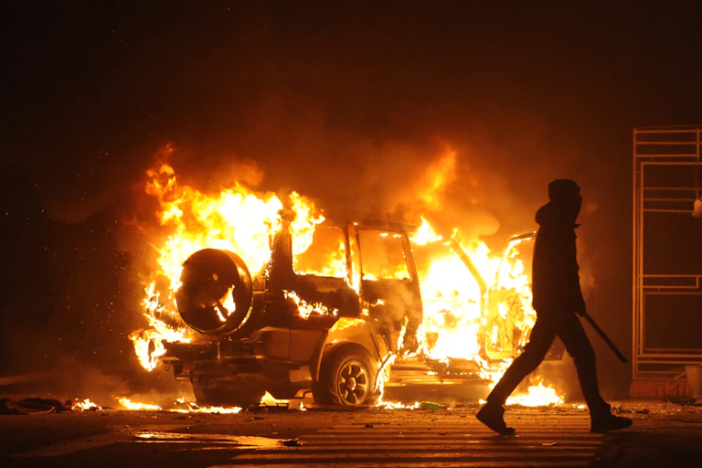 A burning car seen at night with a silhouetted person in a hoodie in the foreground.