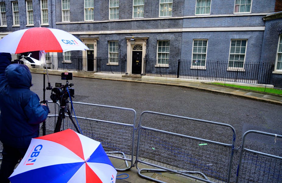 On a rainy day in the press pen across from Number 10 Downing Street, reporters hold red white and blue umbrellas with the GB News logo