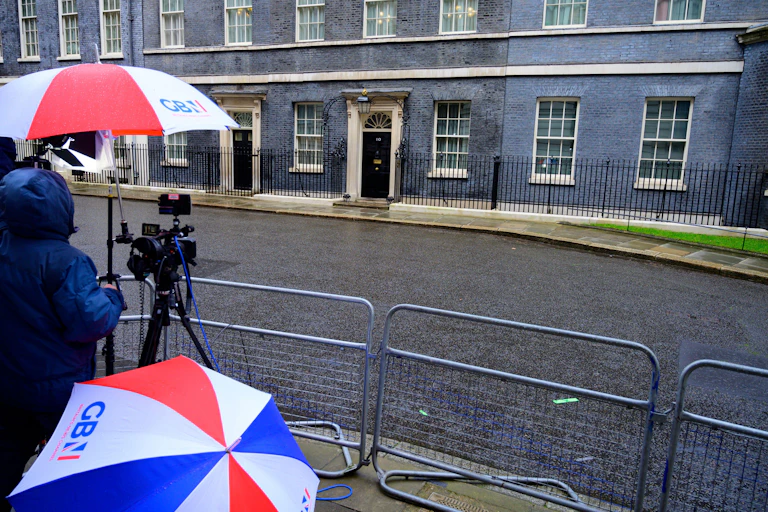 On a rainy day in the press pen across from Number 10 Downing Street, reporters hold red white and blue umbrellas with the GB News logo
