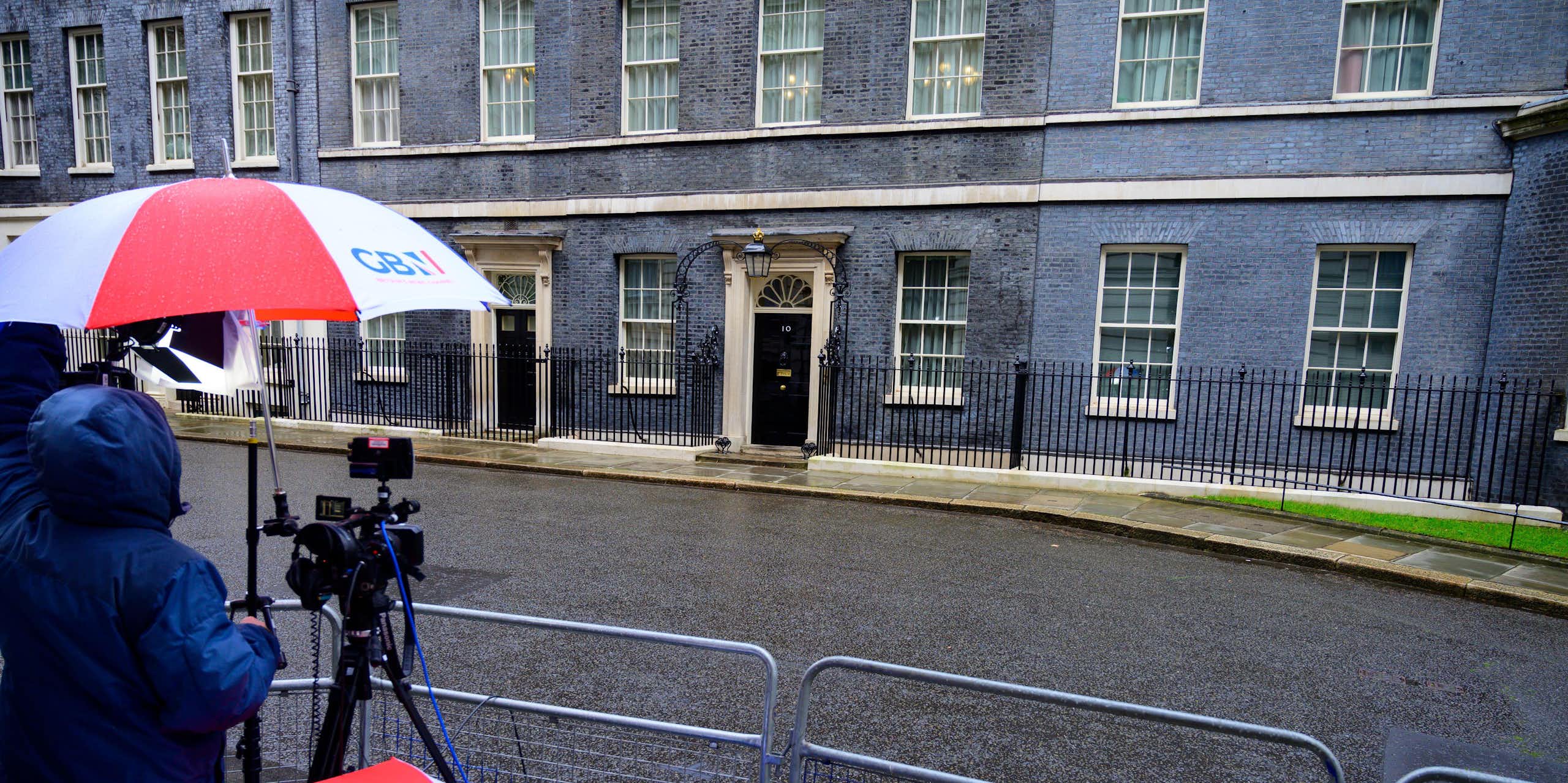 On a rainy day in the press pen across from Number 10 Downing Street, reporters hold red white and blue umbrellas with the GB News logo