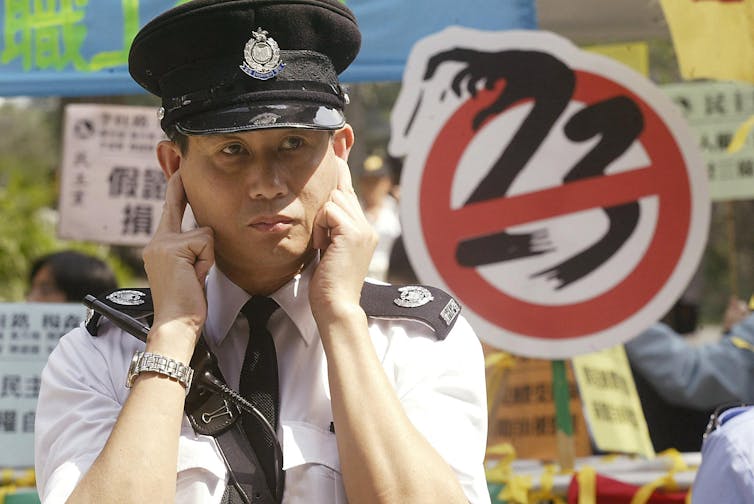 A uniformed police officer puts his fingers in his ears in front of a sign that has the number 23 crossed out.