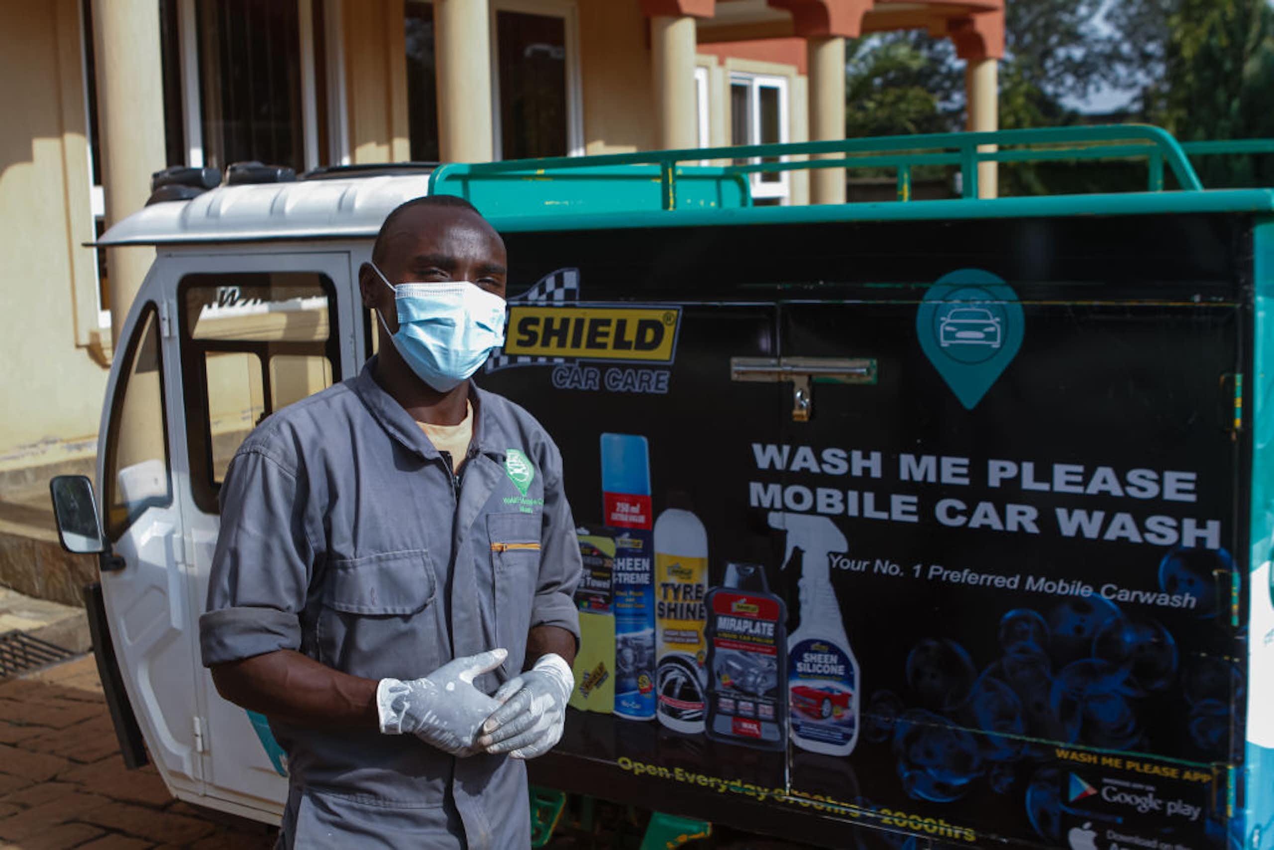 A young man in face mask and disposable gloves stands in front of a vehicle with car wash signage