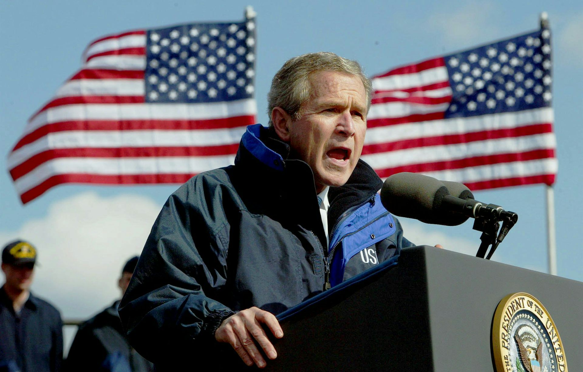 A man speaks into a microphone with two american flags flying behind him