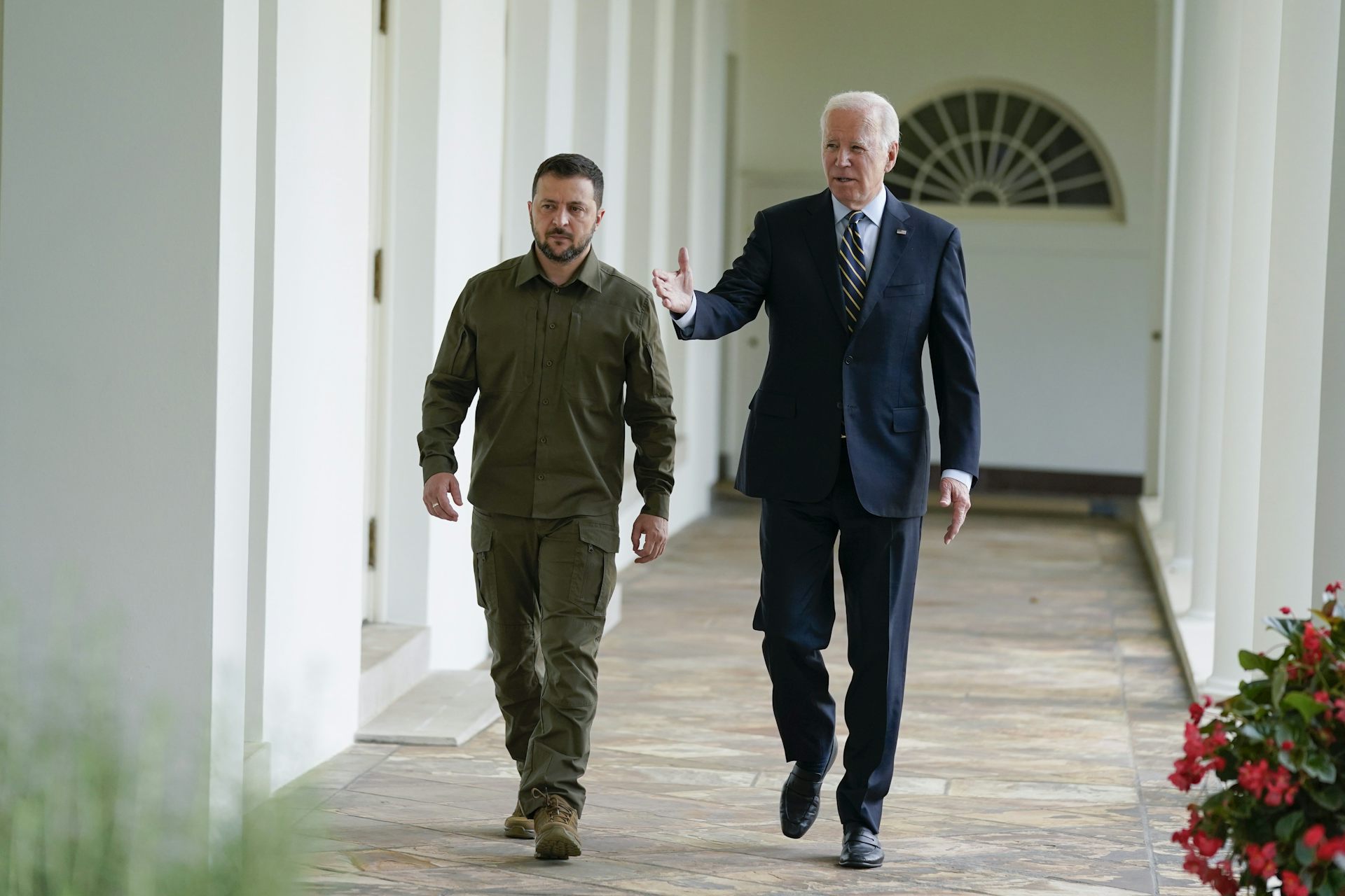 An older man in a suit walks with a younger man in dark green military clothing along a white stone colonnade.