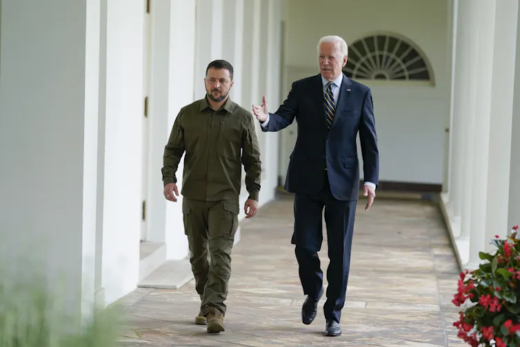 An older man in a suit walks with a younger man in dark green military clothing along a white stone colonnade.