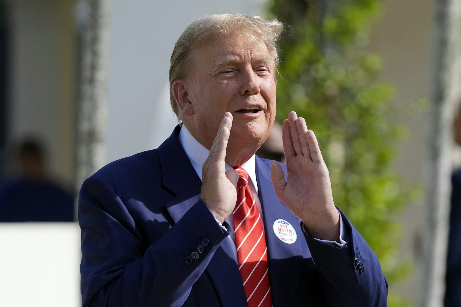A rotund man with greyish blond hair in a blue suit and red tie holds his hands on either side of his mouth as he speaks.